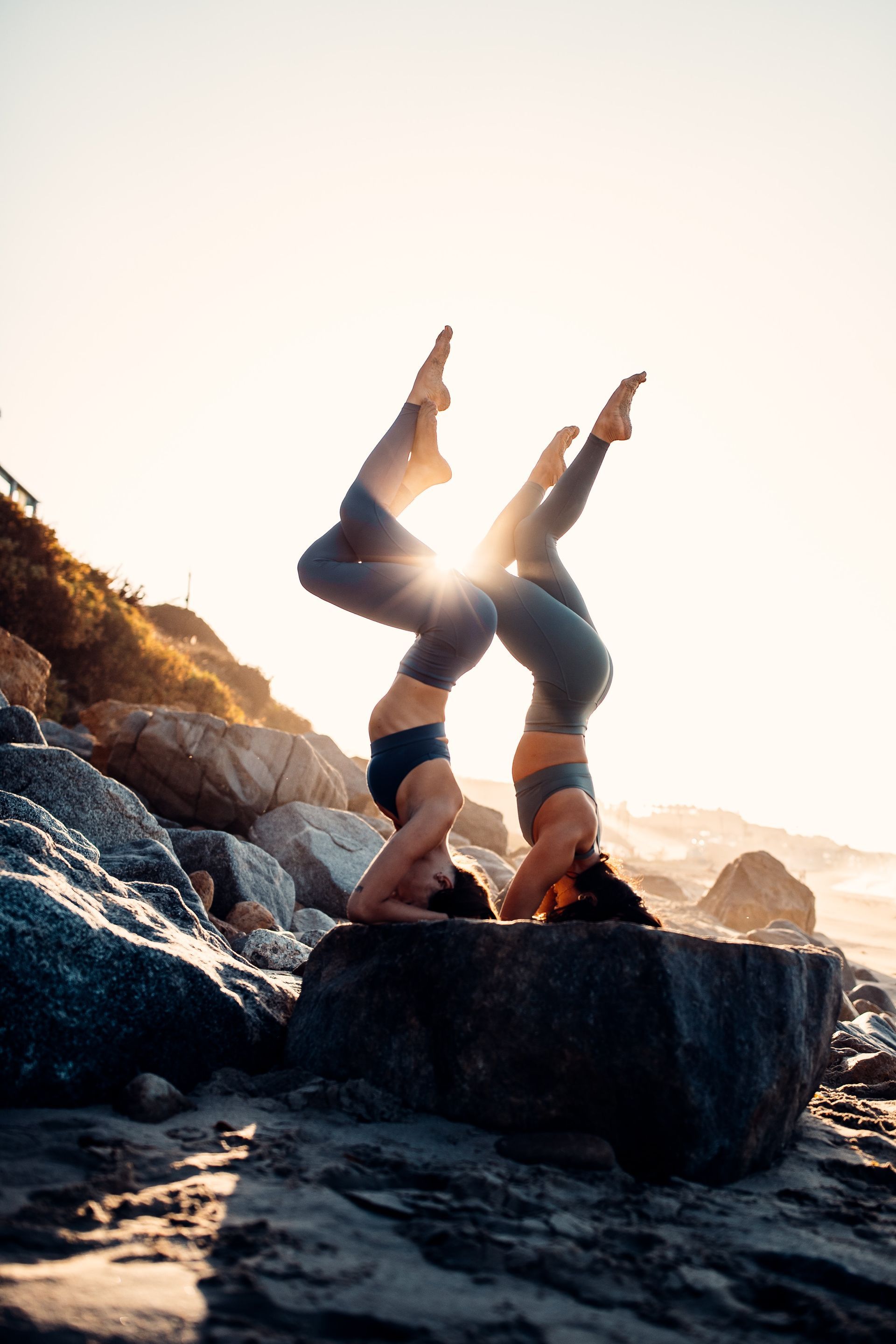 Two people doing headstand yoga on a rock, legs intertwined, against a sunny beach background.