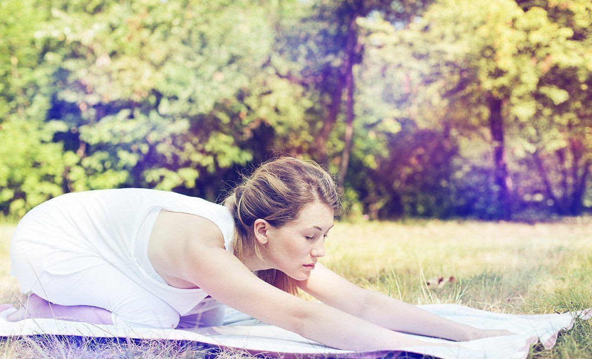 A woman in a white dress is stretching on a yoga mat in the grass.