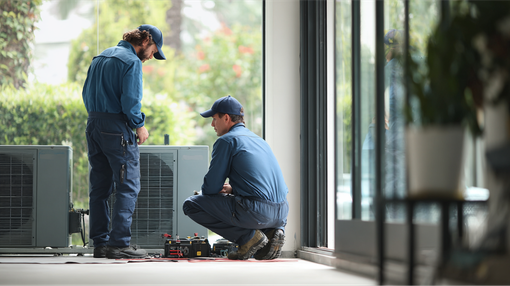 Two workers in blue coveralls inspect an air conditioning unit near a large window.