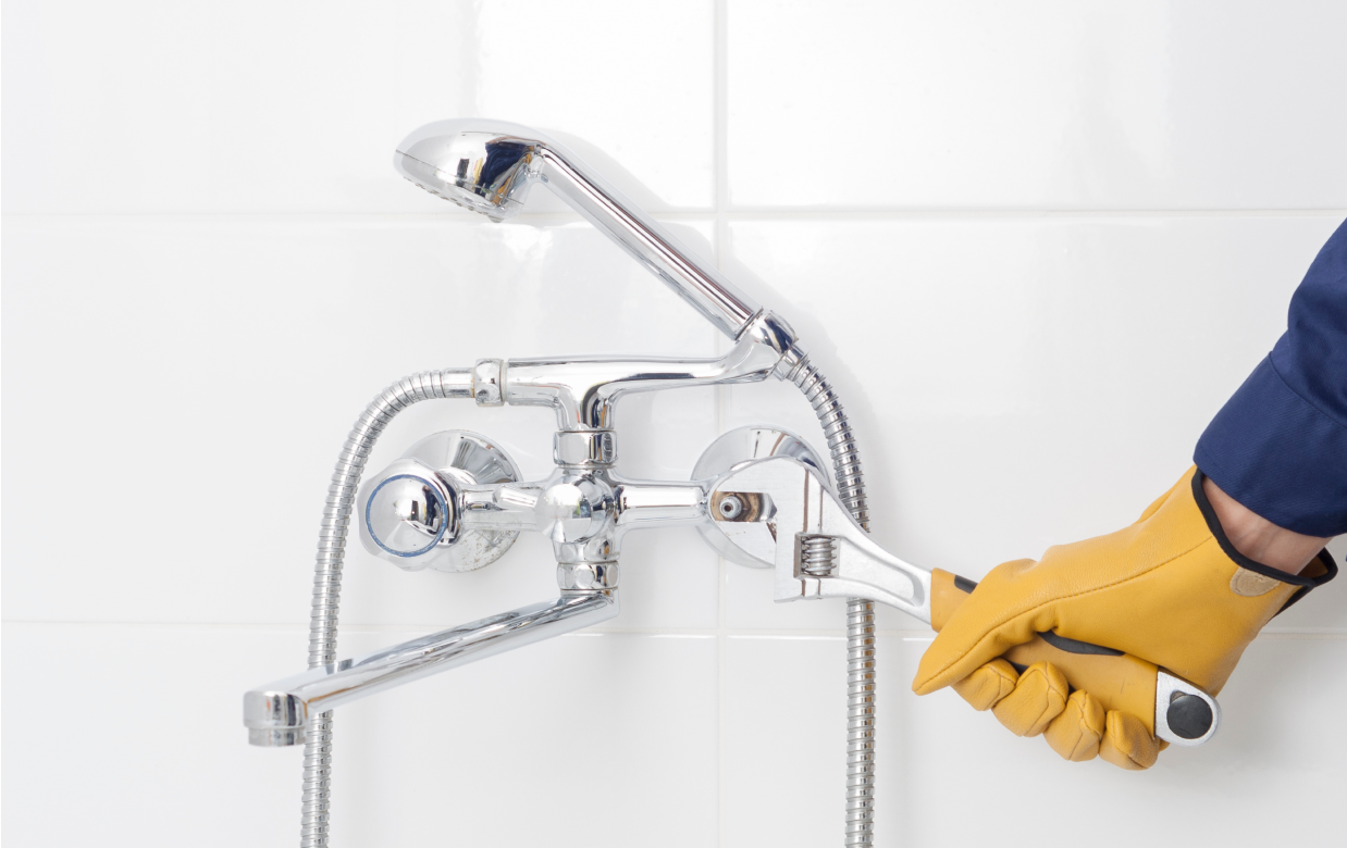 Hand in yellow glove using wrench to fix a shower faucet in a tiled bathroom.