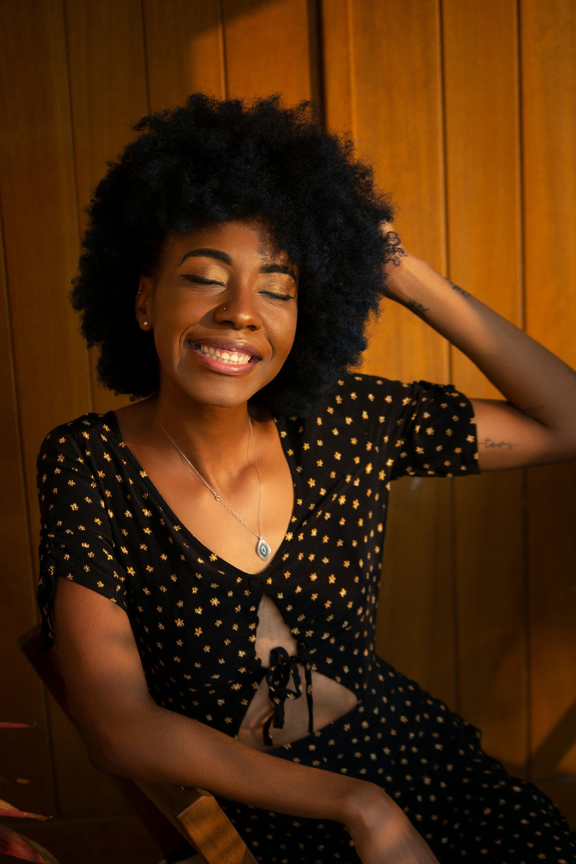 A woman in a black polka dot dress is smiling while sitting in front of a wooden wall.