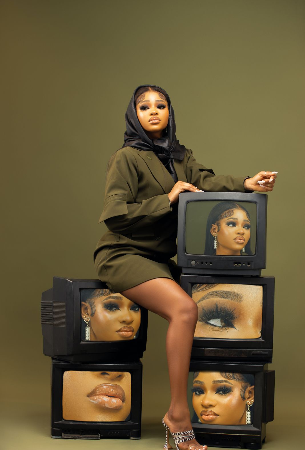 A woman is sitting on a stack of televisions with her face on them.