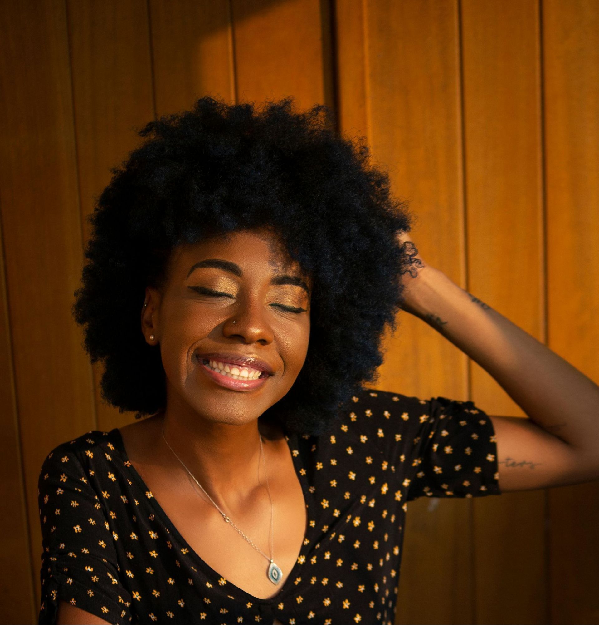 Woman with dark, voluminous hair smiles, eyes closed. Wearing a black polka-dot top, necklace, arm raised in sunlight.