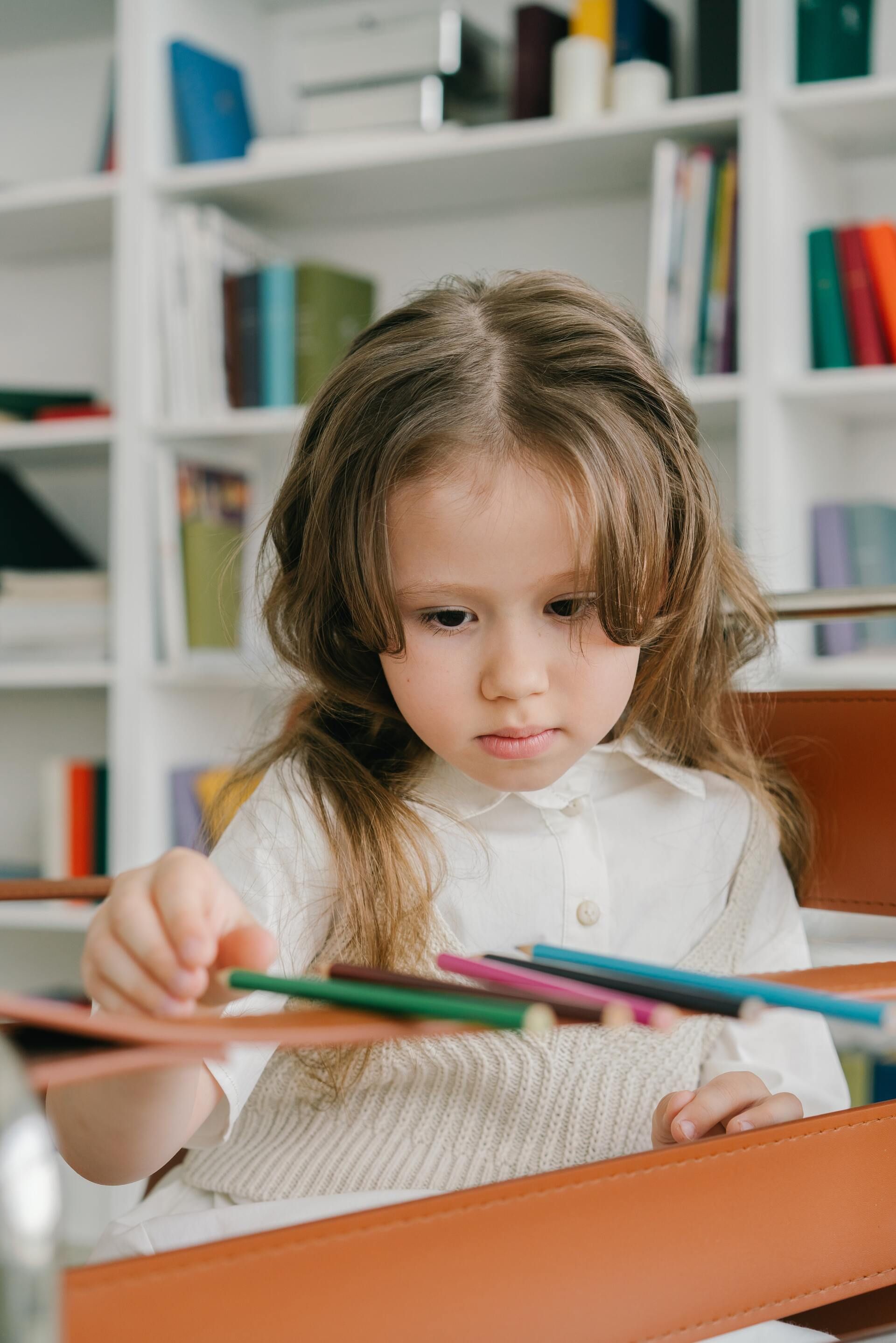 A little girl is sitting at a table holding a bunch of colored pencils.