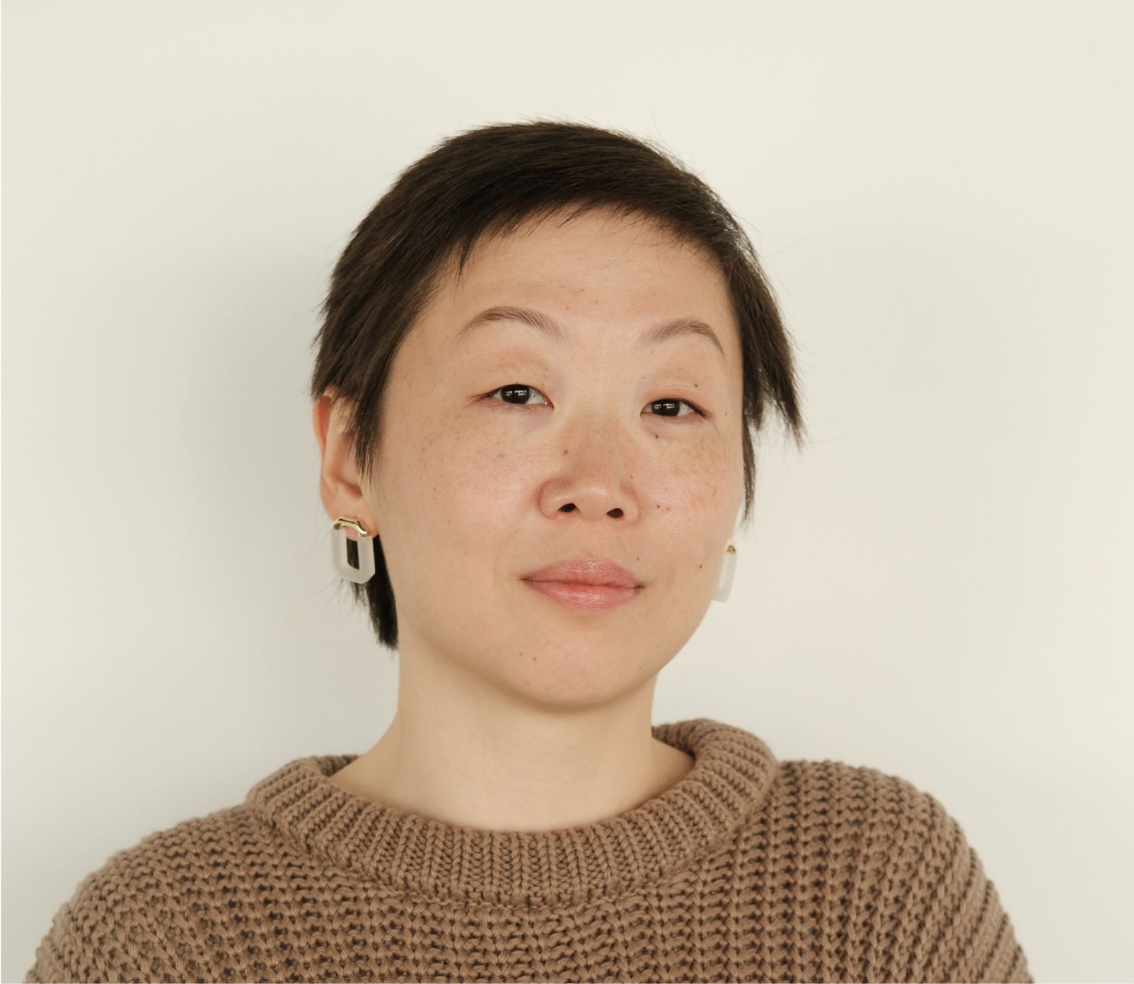 A woman wearing a brown sweater and earrings looks at the camera