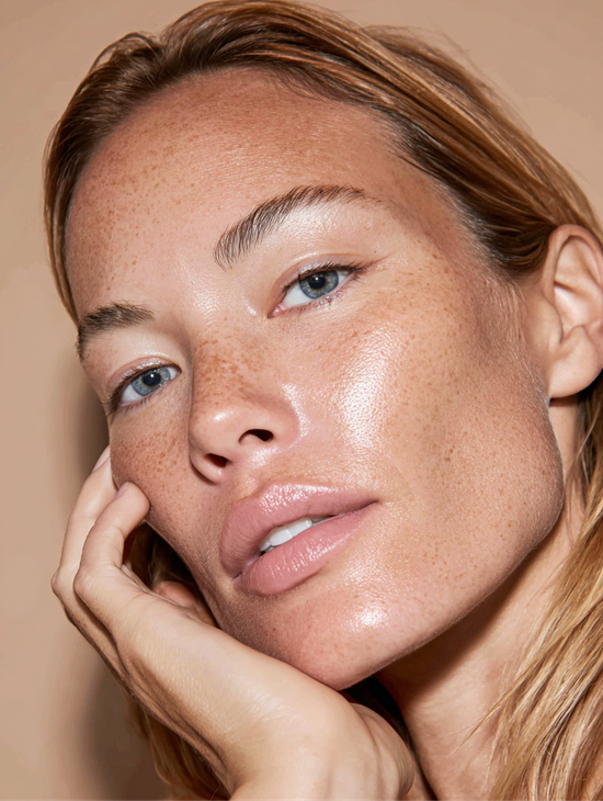 Woman with freckled, dewy skin, gazing at the camera, hand touching face; neutral background.