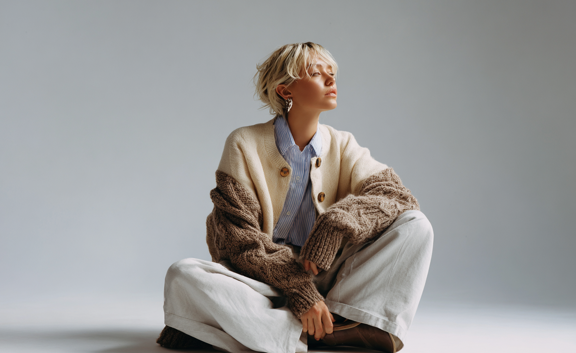 Person with short blonde hair sits cross-legged, wearing a cardigan, striped shirt