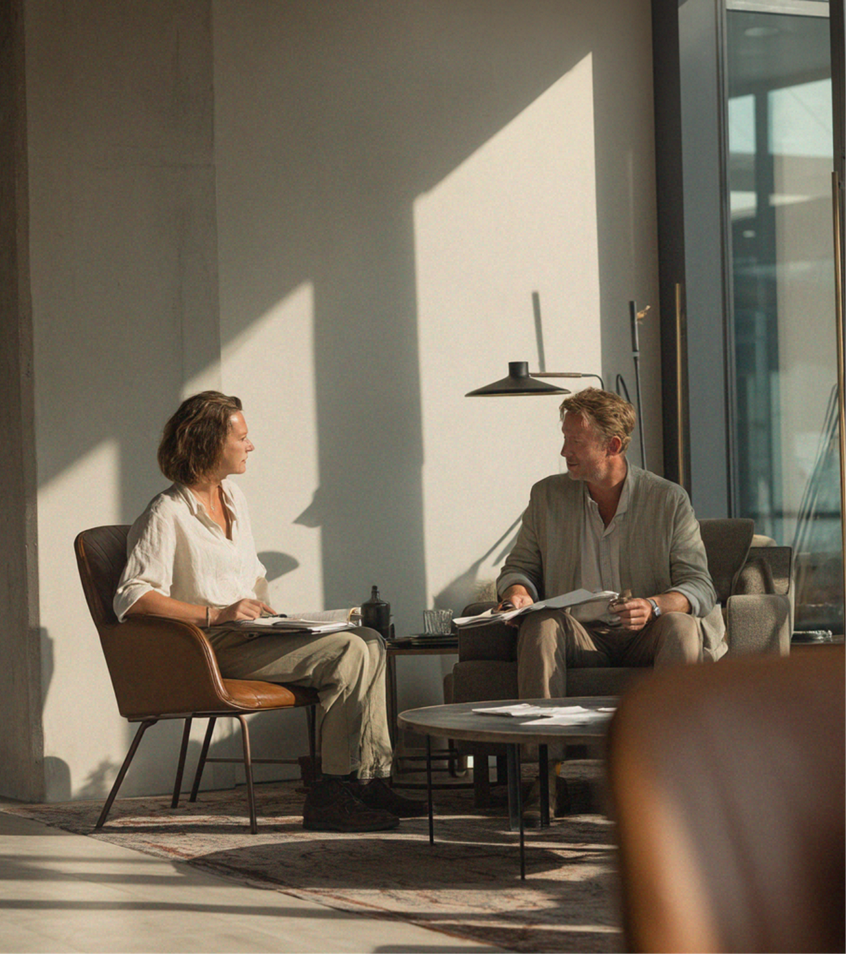Two people chatting in a sunlit modern living room, seated by a window and coffee table.