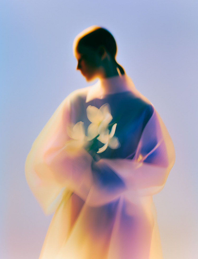 A soft-focus, ethereal portrait of a person holding white flowers against a gradient background of blue and warm light.