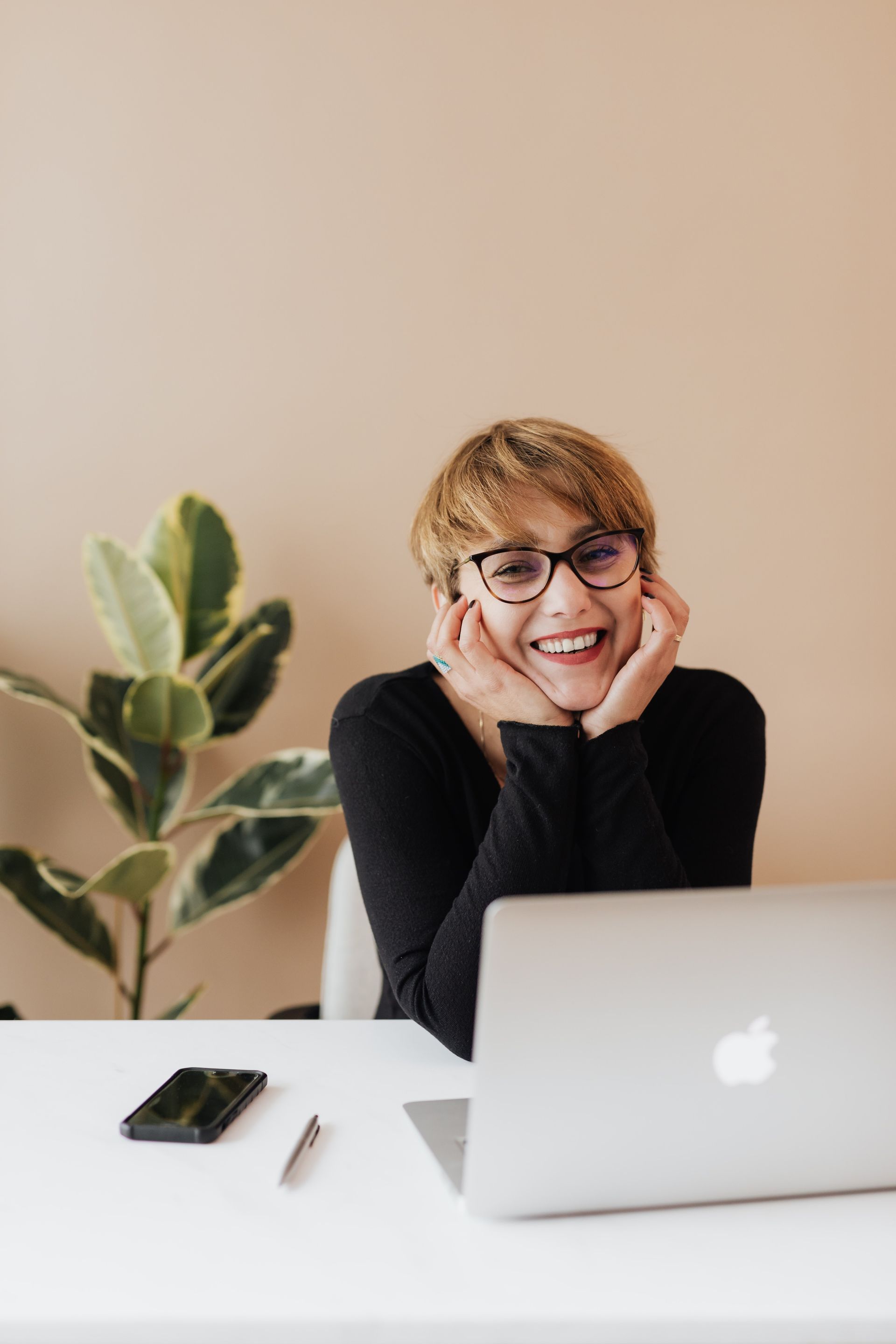 Mujer sentada en un escritorio con una laptop, sonriendo, con la barbilla apoyada en las manos. Interior con una planta.
