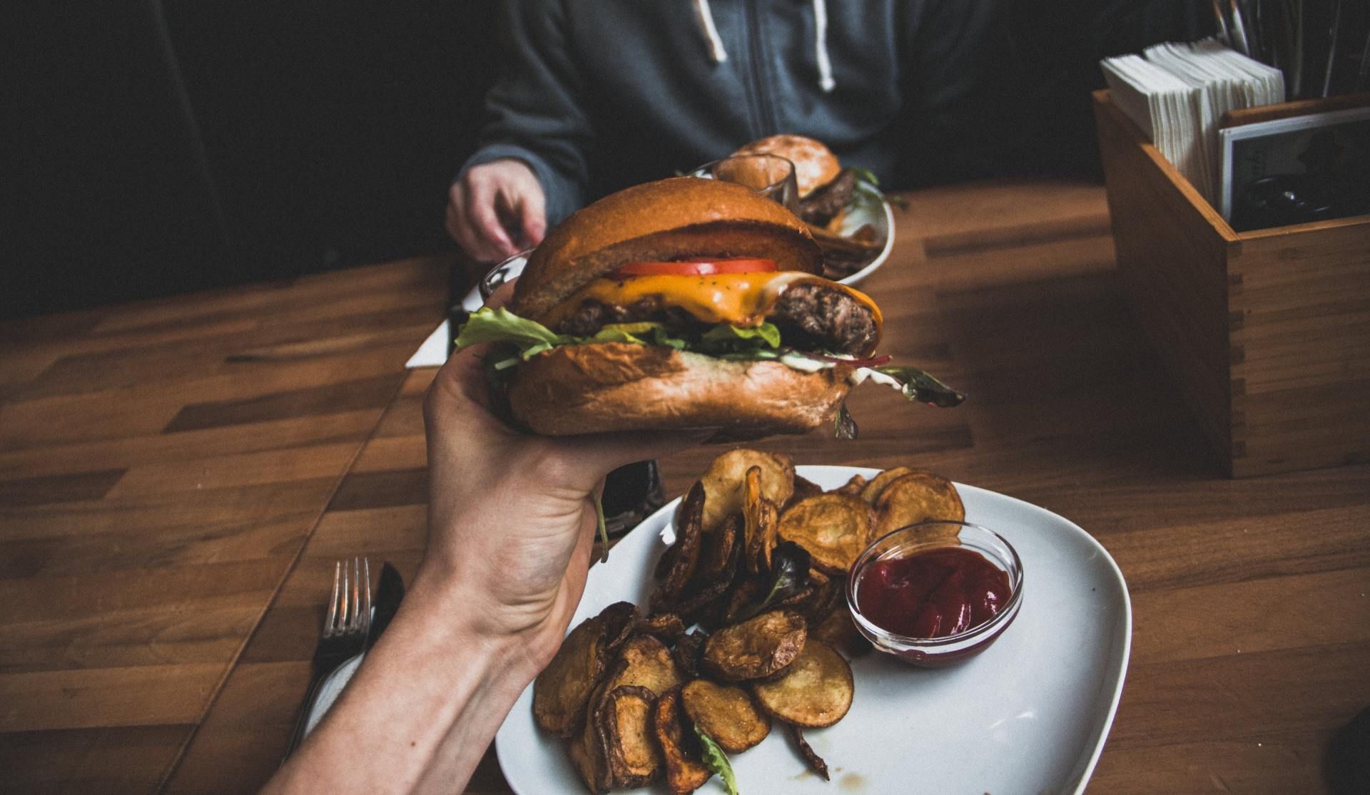 A person is holding a hamburger over a plate of potato chips.