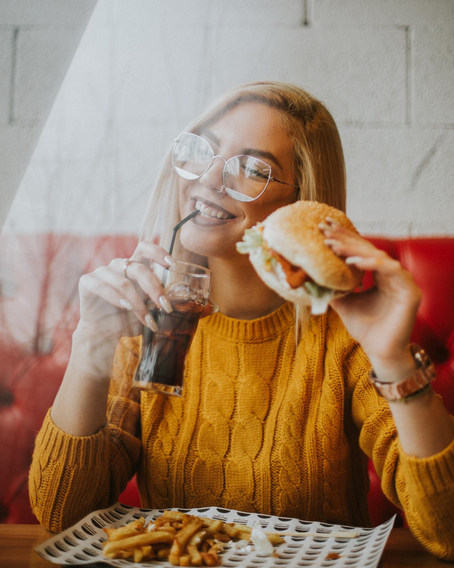 A woman in a yellow sweater is eating a hamburger and drinking a soda.
