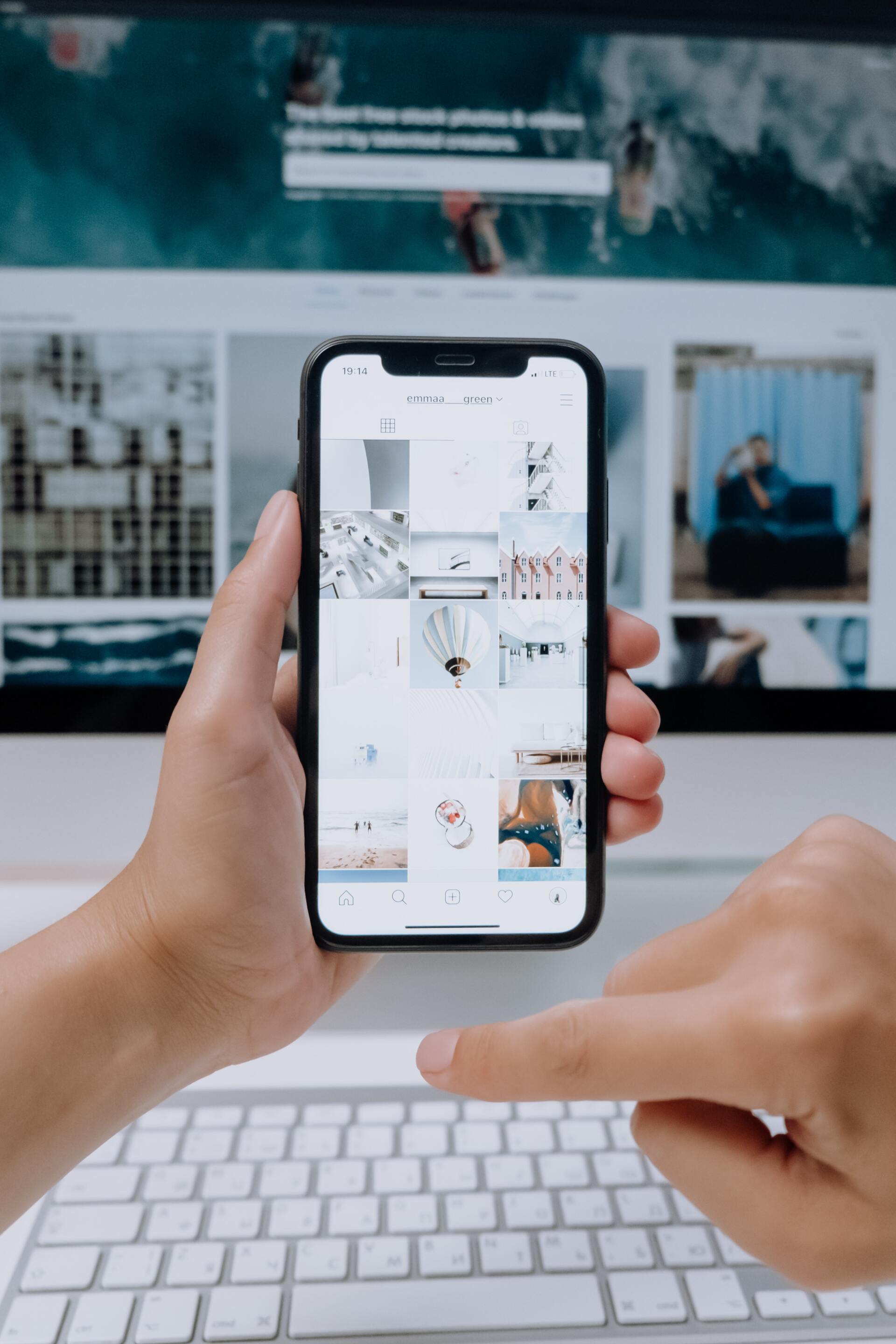 Person holding a phone displaying an Instagram feed, with a computer and keyboard in the background.