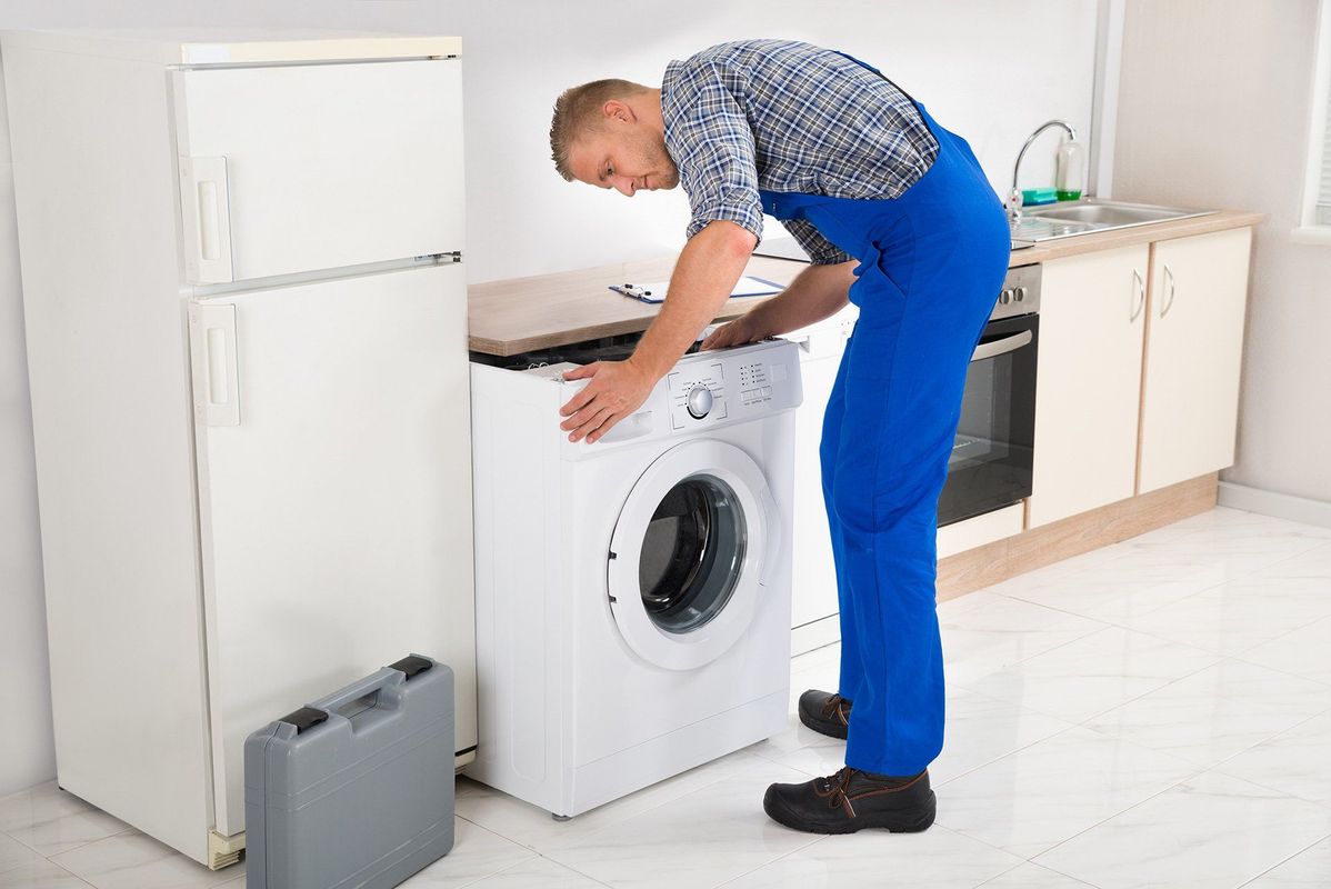 A man in blue overalls is working on a washing machine in a kitchen.