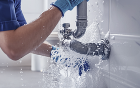 Plumber in blue gloves fixing a leaking pipe in a bathroom, water spraying.
