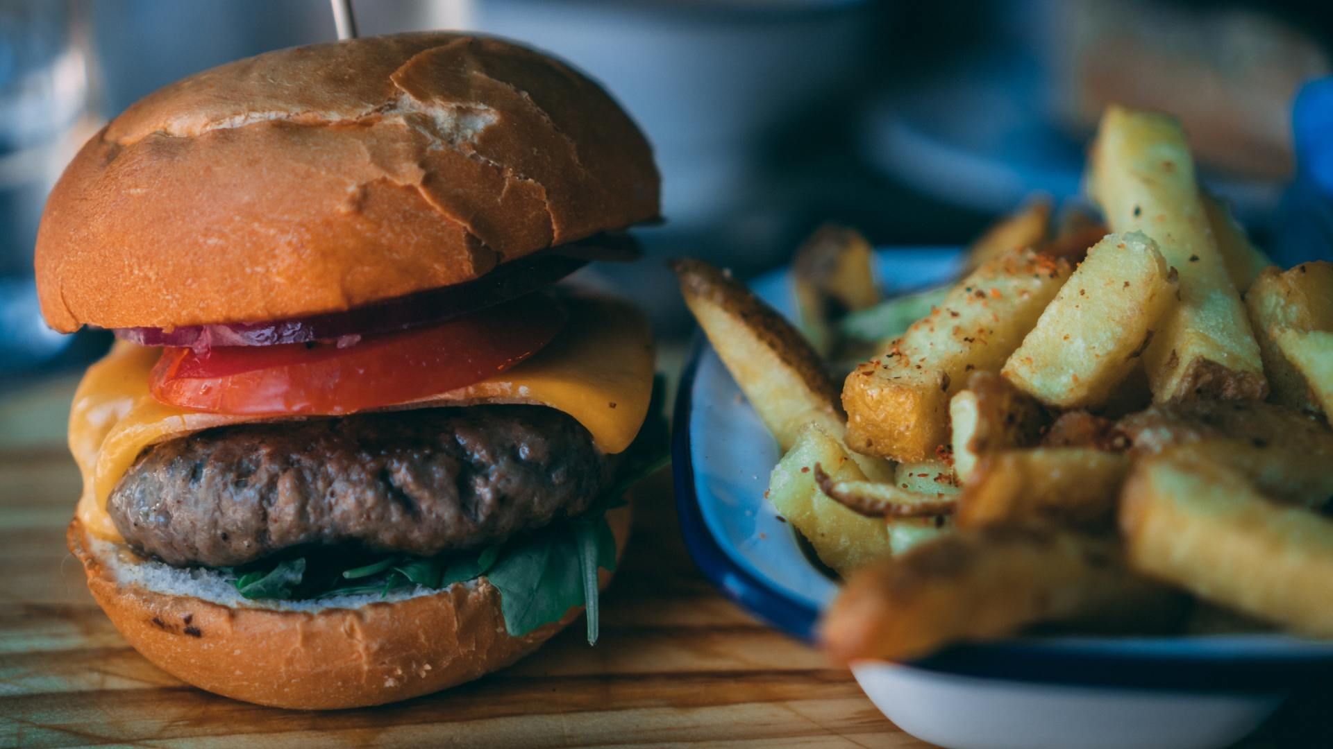 A juicy cheeseburger with a tomato slice and greens next to a plate of thick-cut, golden-brown fries on a wooden surface.