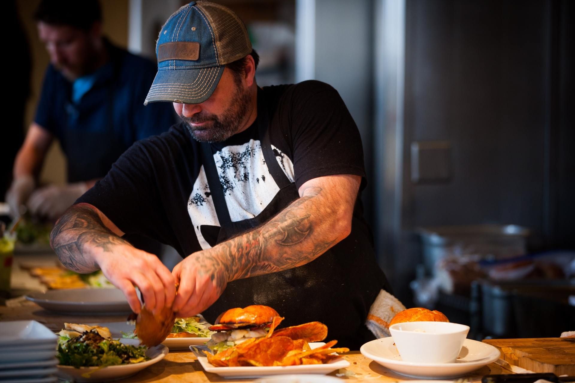 A chef wearing a baseball cap and apron prepares food on plates in a professional kitchen.