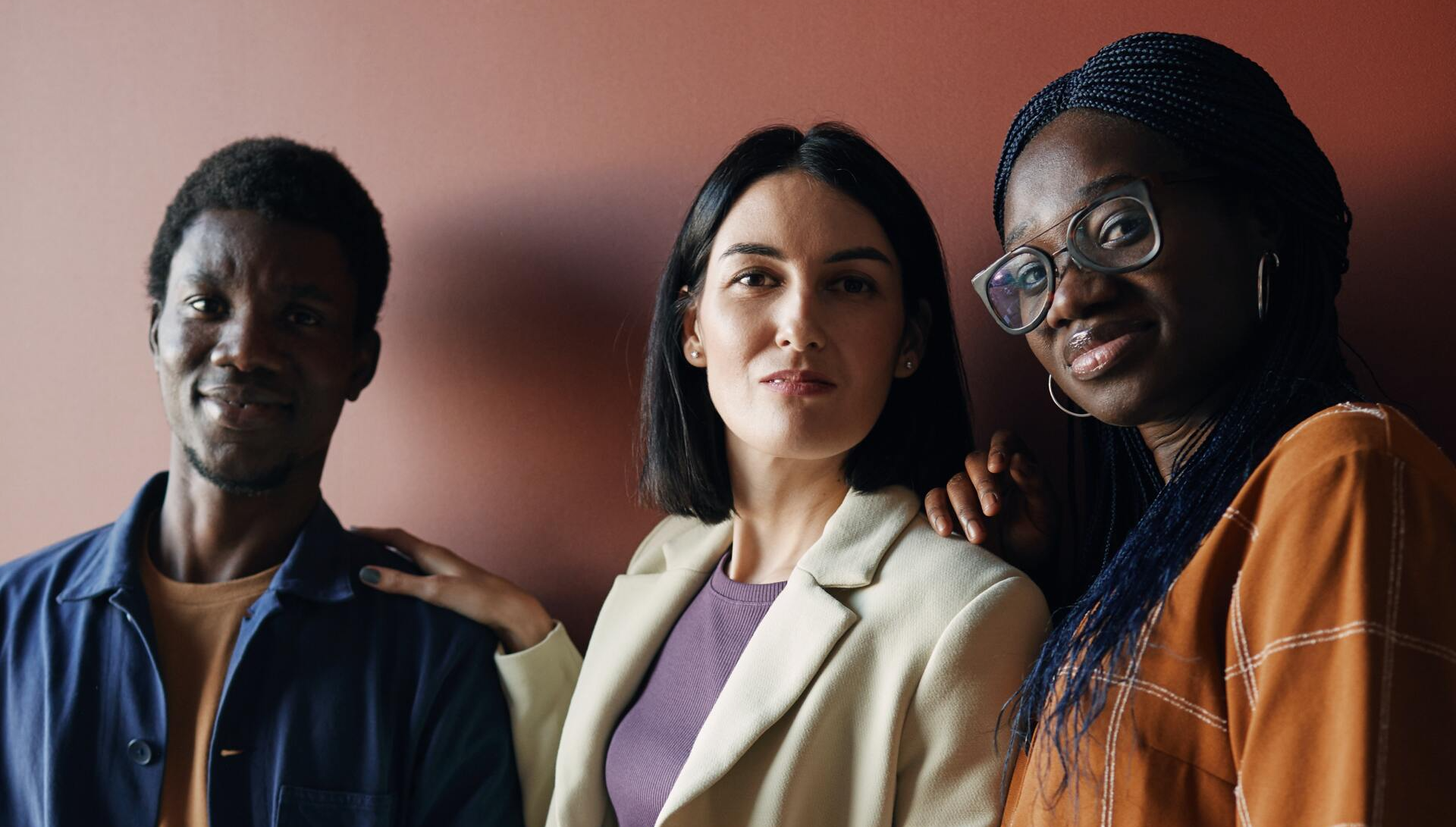 Three people posing closely, the middle person's arm on the first person's shoulder. Neutral wall background.