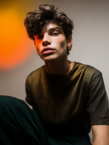 A portrait of a person with curly dark hair wearing a metallic bronze-colored shirt, set against a warm, orange-lit backdrop.