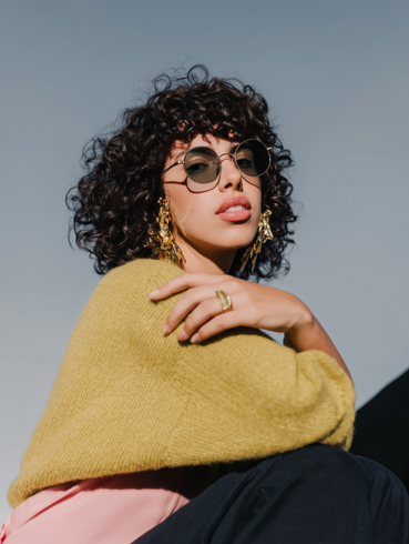 A person with curly dark hair wearing sunglasses, a yellow sweater, and large gold earrings against a blue sky.