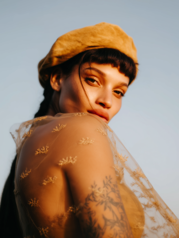 A person with a tattoo on their arm wears a mustard-yellow beret and a sheer, embroidered garment against a blue sky.