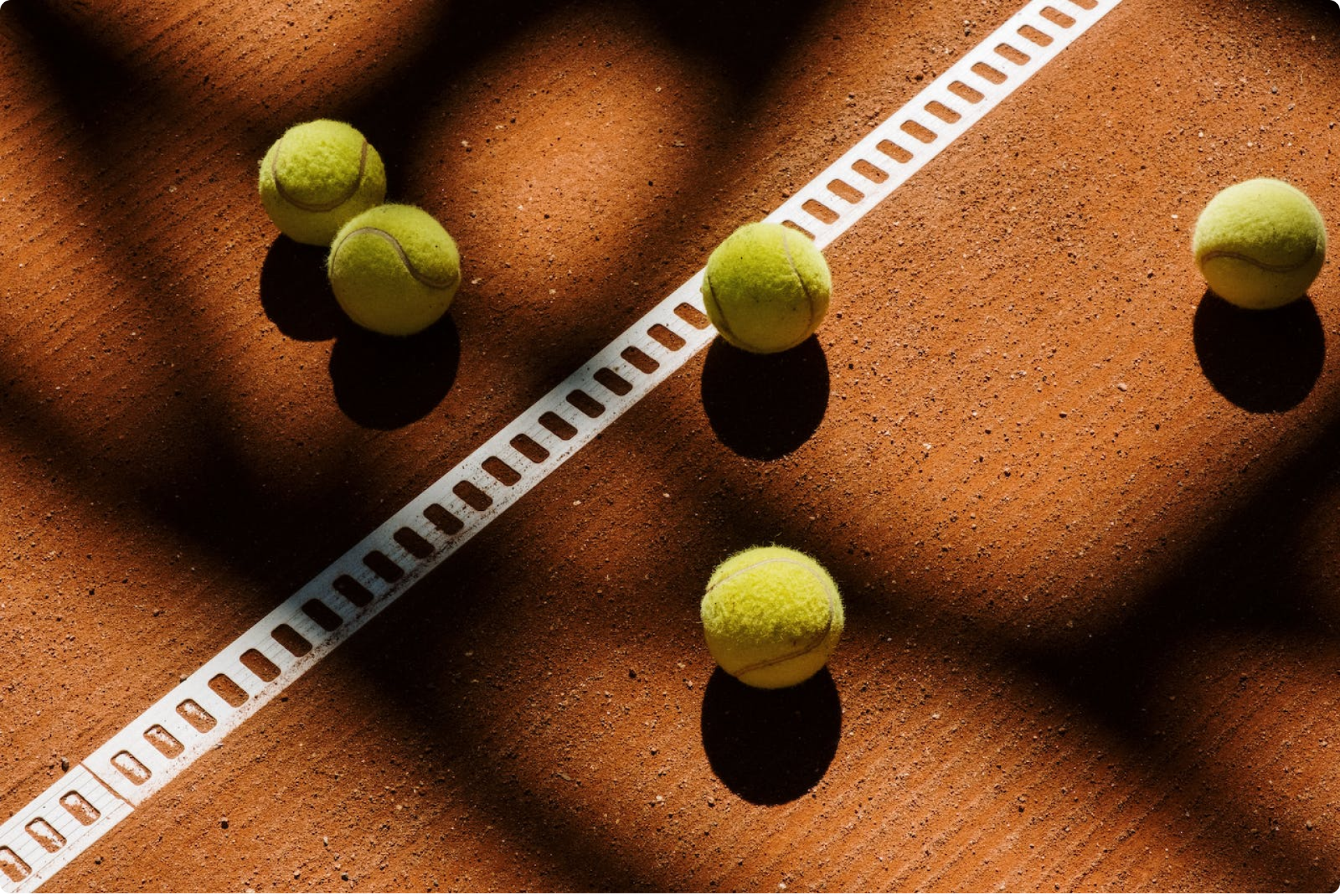 Five yellow tennis balls on a clay court, with a white line crossing diagonally.