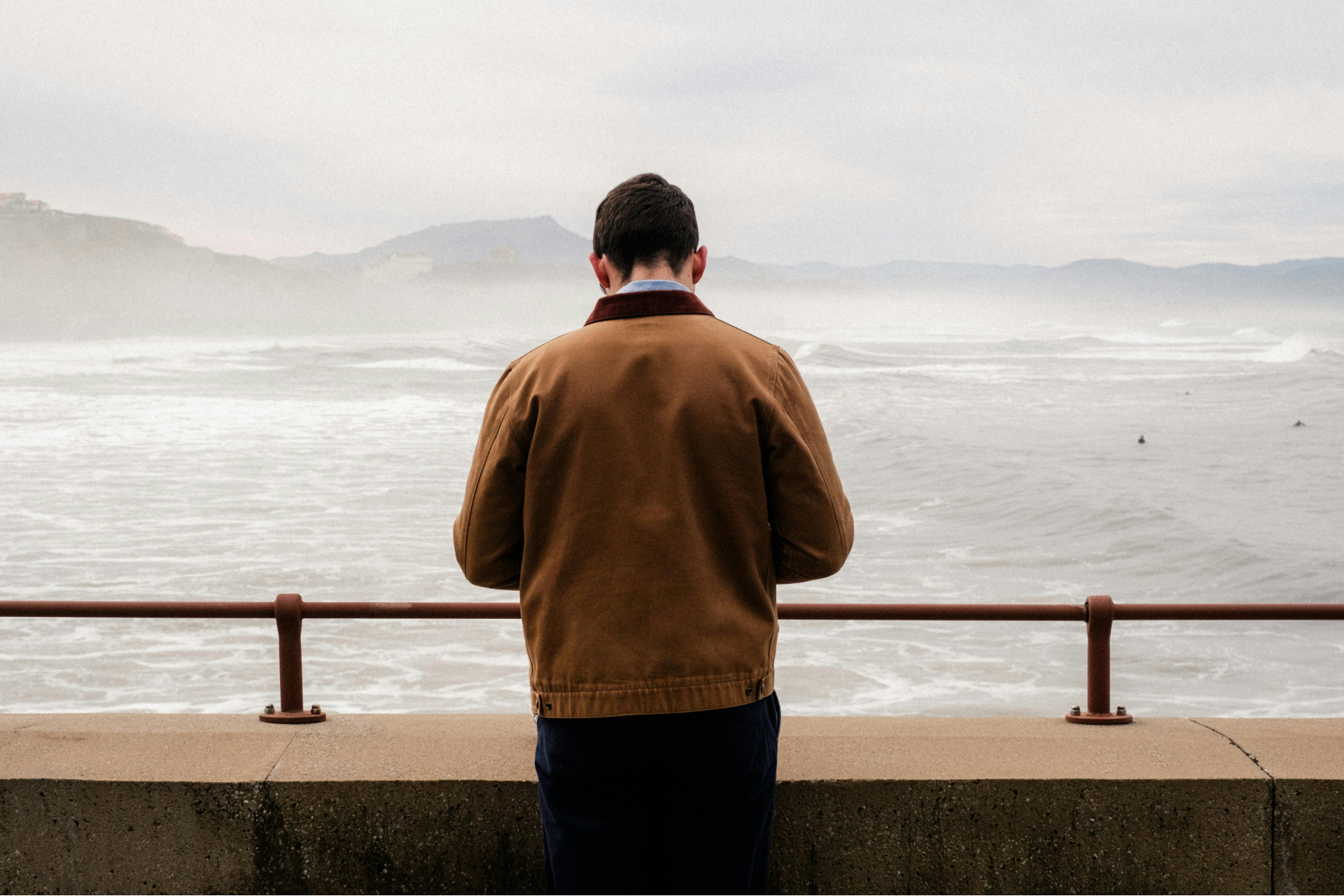 Man in brown jacket looking out at a wavy ocean from behind a concrete barrier with a railing. Overcast sky.