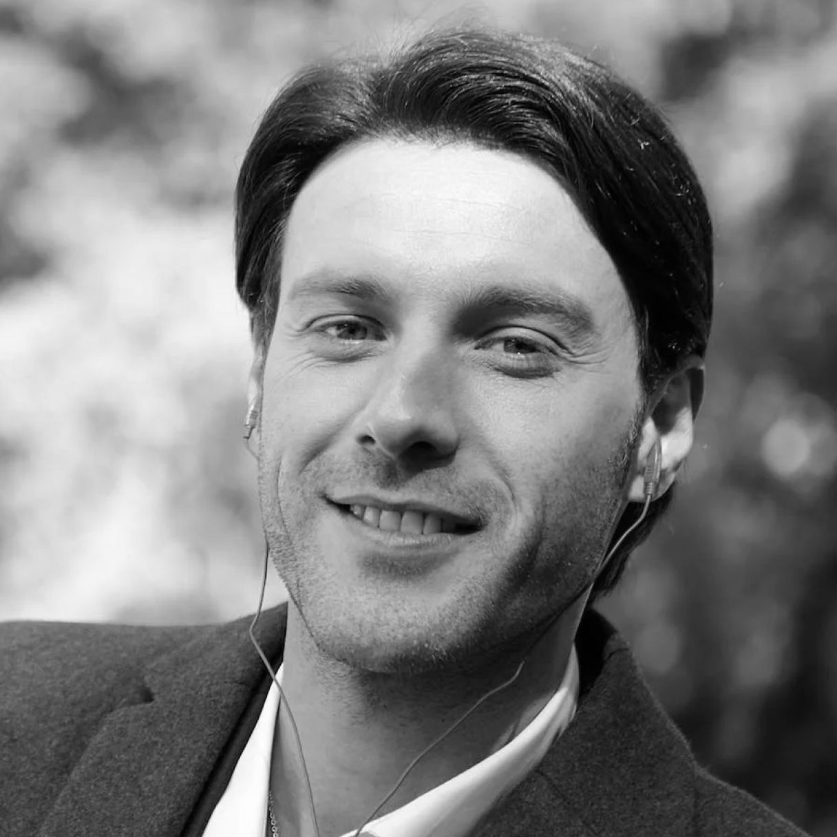 Black-and-white portrait of a smiling person wearing headphones and a blazer against a blurred outdoor background.