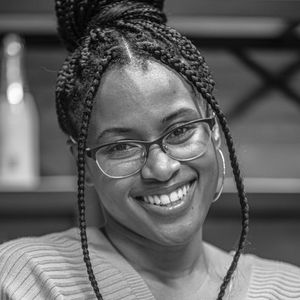 Black and white portrait of a smiling Black woman with glasses and braided hair, indoors.