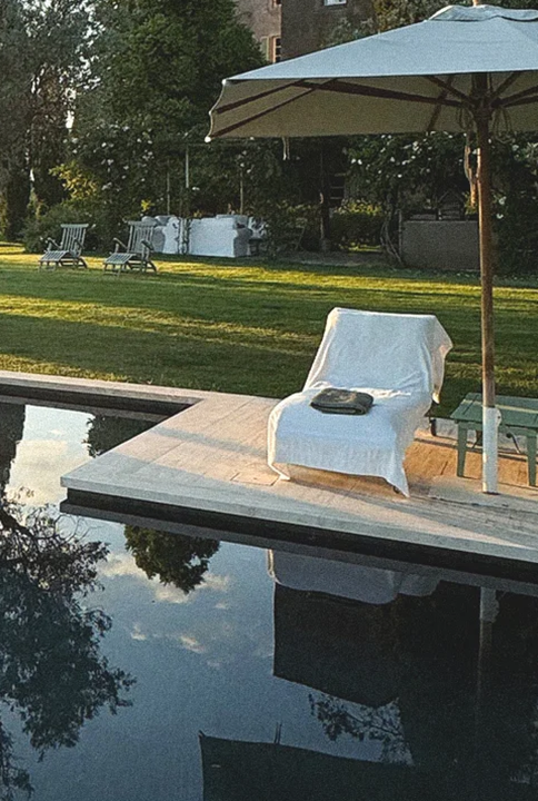 Una sdraio bianca sul bordo piscina, accanto a un ombrellone, con un prato e degli alberi sullo sfondo.
