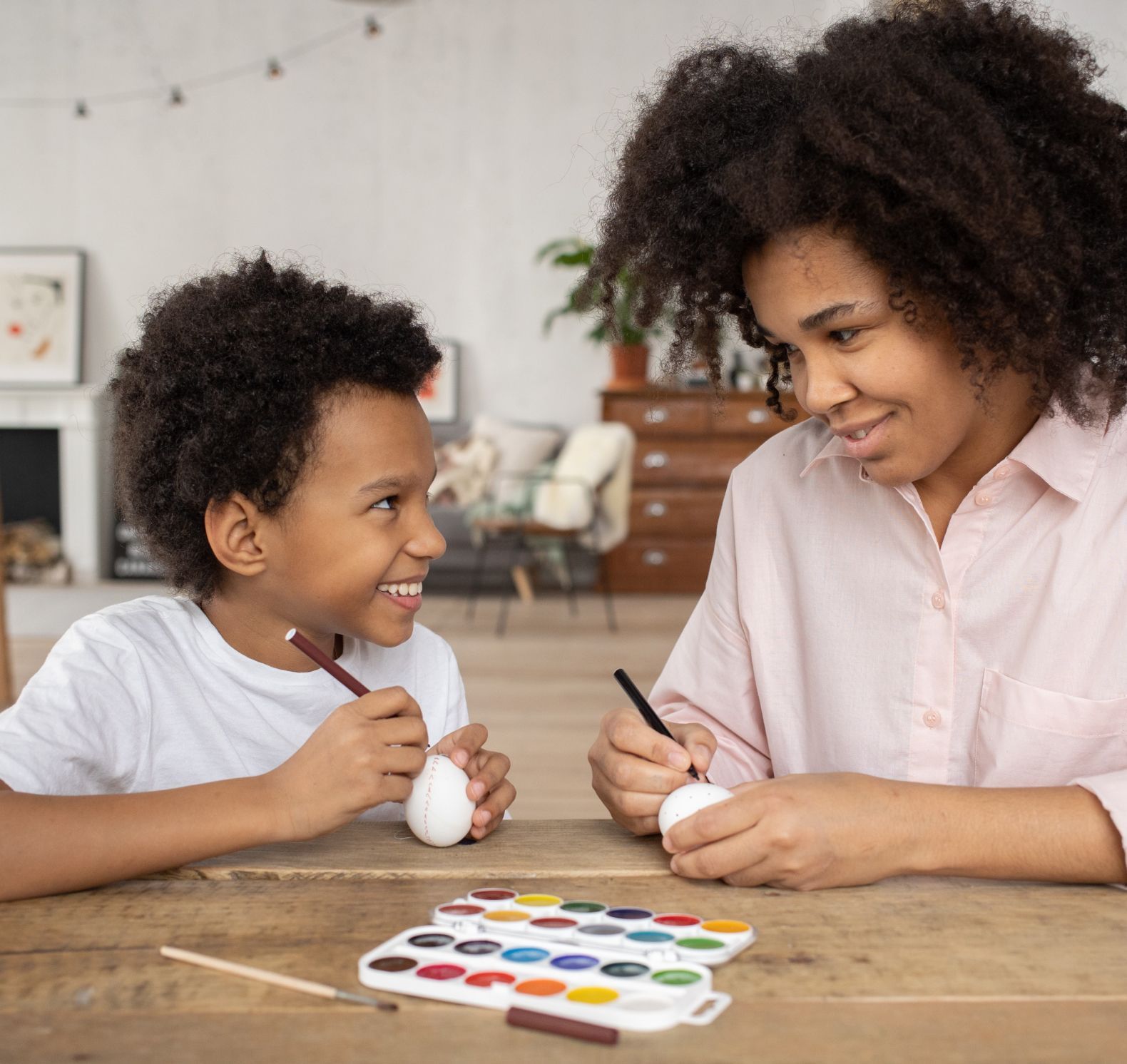 A woman and child happily decorate Easter eggs together at a wooden table. A watercolor palette is visible.