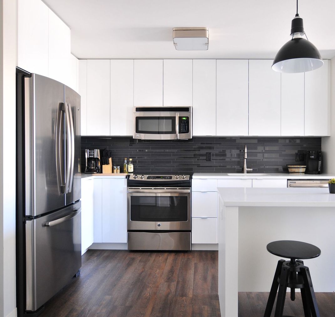 Cuisine blanche moderne avec électroménagers en inox, parquet en bois foncé et crédence noire. Une suspension noire est suspendue au-dessus d'un plan de travail blanc avec tabouret.