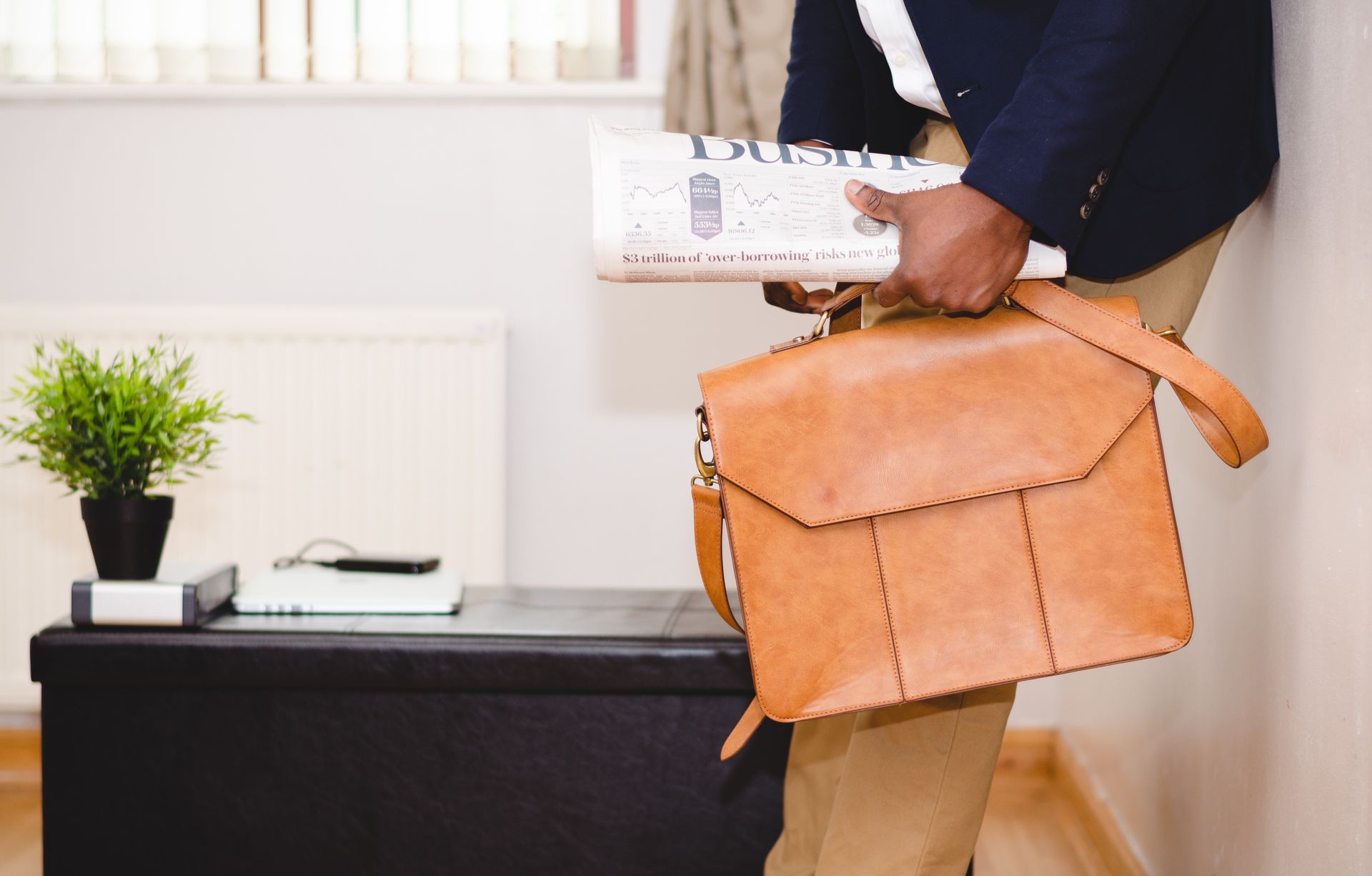 Person holding a newspaper and a leather briefcase, leaning against a wall. There is a small plant on a dark cabinet.