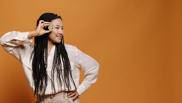 A woman is holding a kiwifruit in front of her eye.