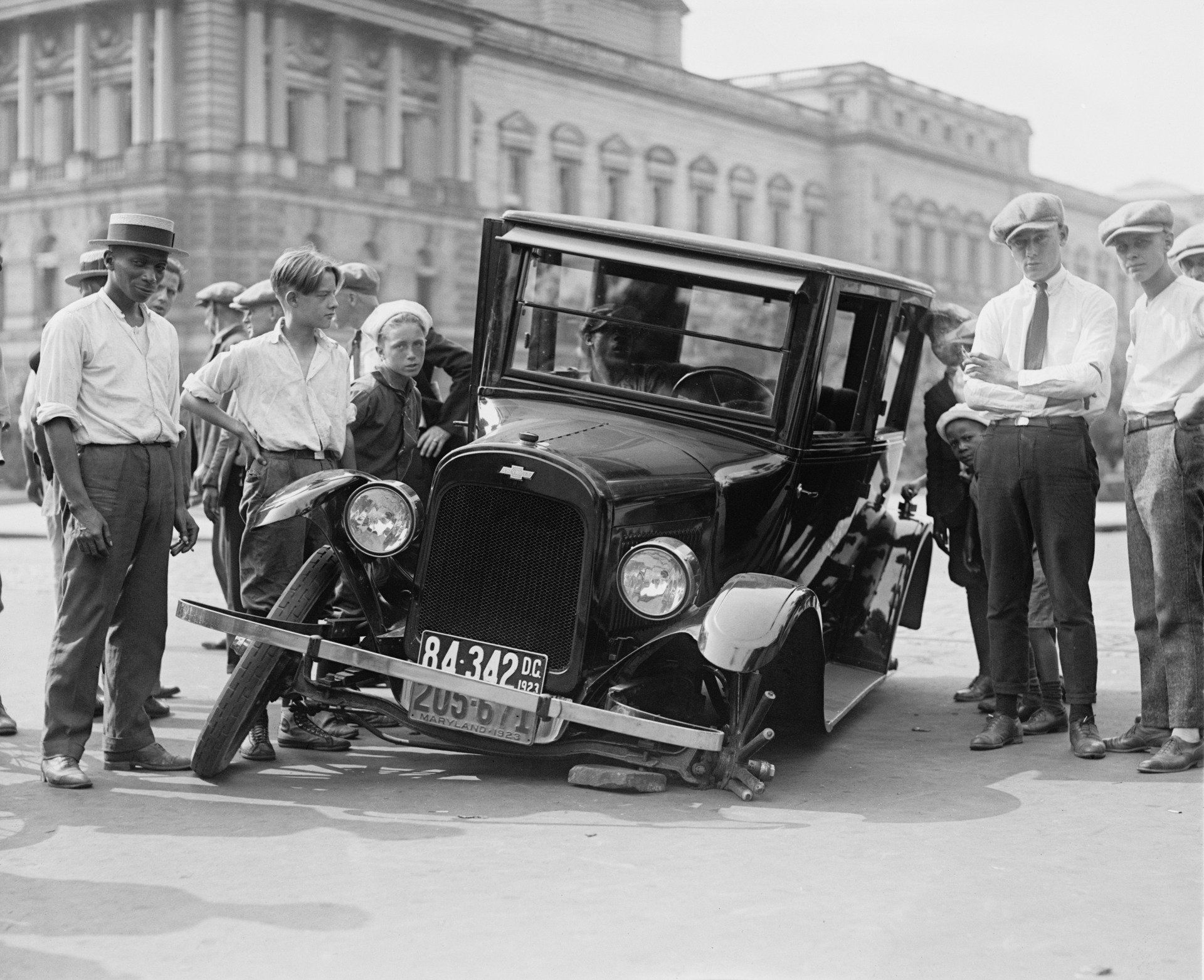 Old time picture showing an old car with its front wheel missing and several boys standing on either side of the vehicle.