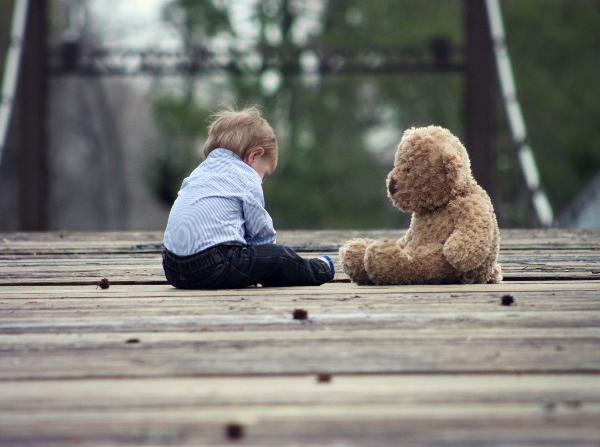 a little boy sits next to a teddy bear on a wooden deck