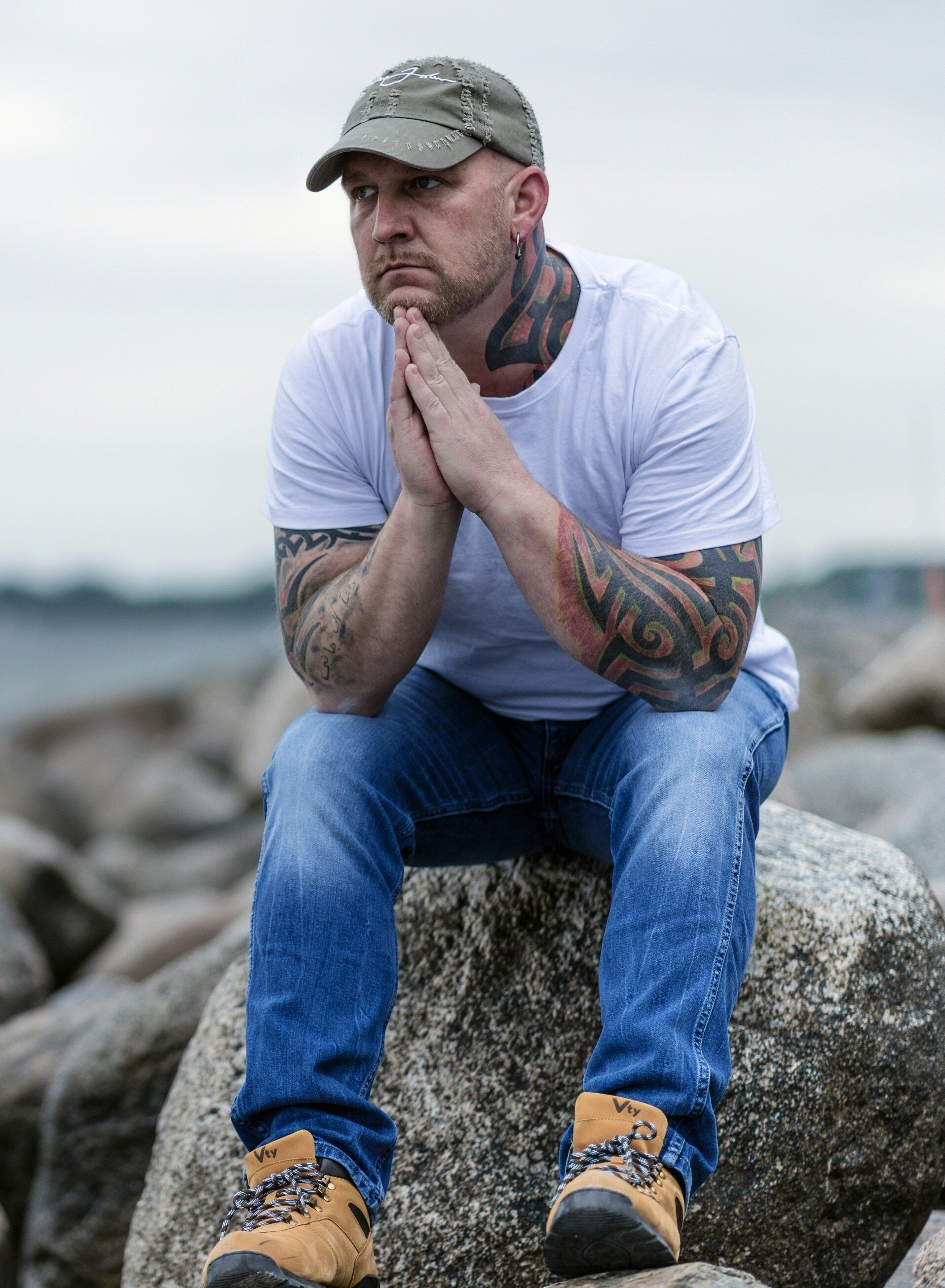 A man with tattoos is sitting on a rock with his hands folded.
