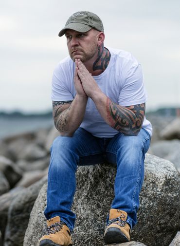 A man with tattoos is sitting on a rock with his hands folded.