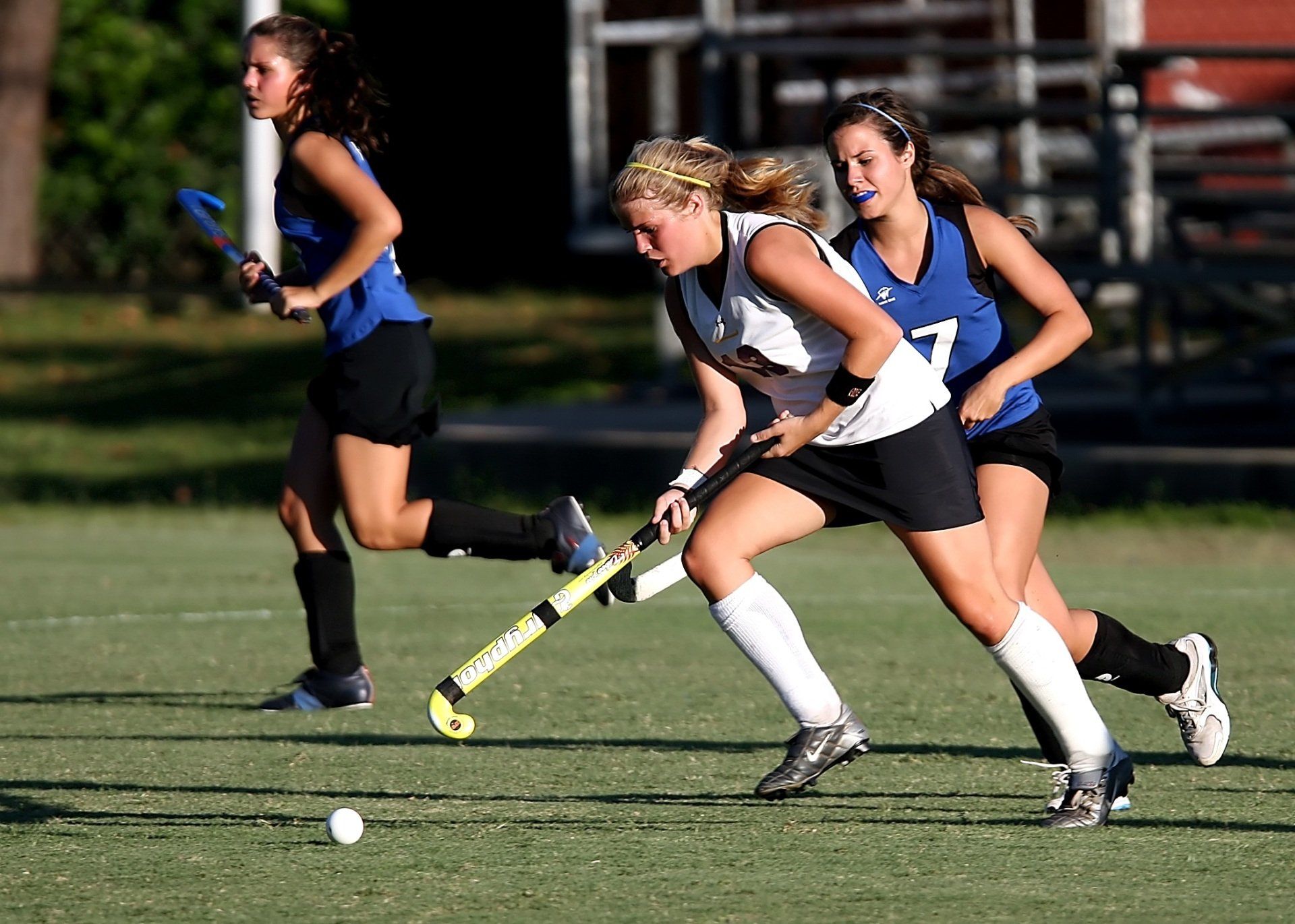 Hockeyspelers op groen gras, één in het wit en zwart, dribbelen met de bal; twee anderen rennen erachteraan.