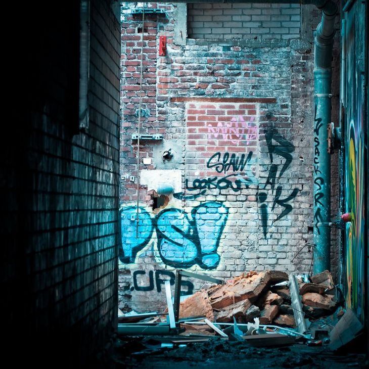 The walls of a brick alley, covered in spray paint, with rubbish strewn all over the ground