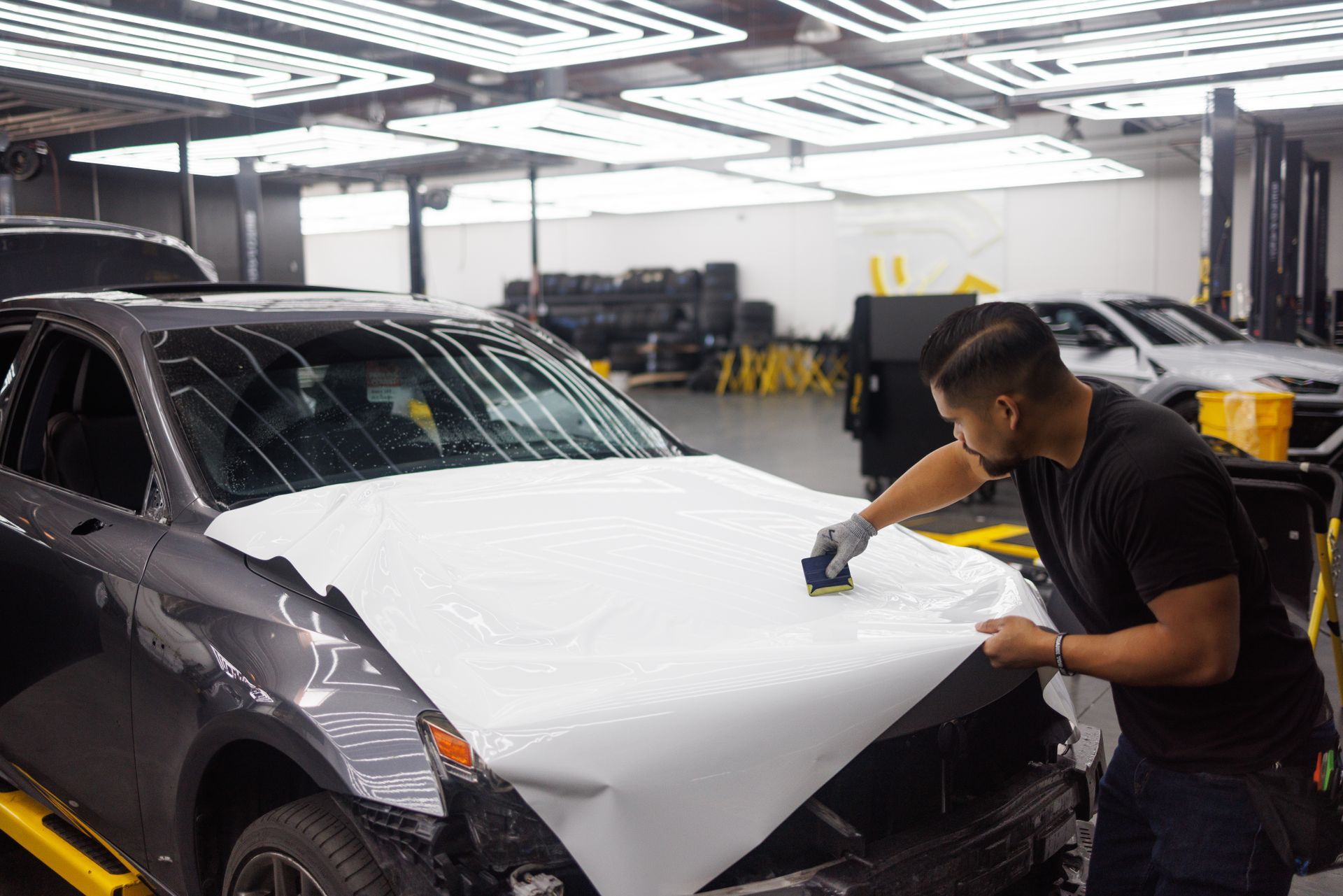 Two people applying white vinyl wrap to a gray car hood inside a well-lit garage.