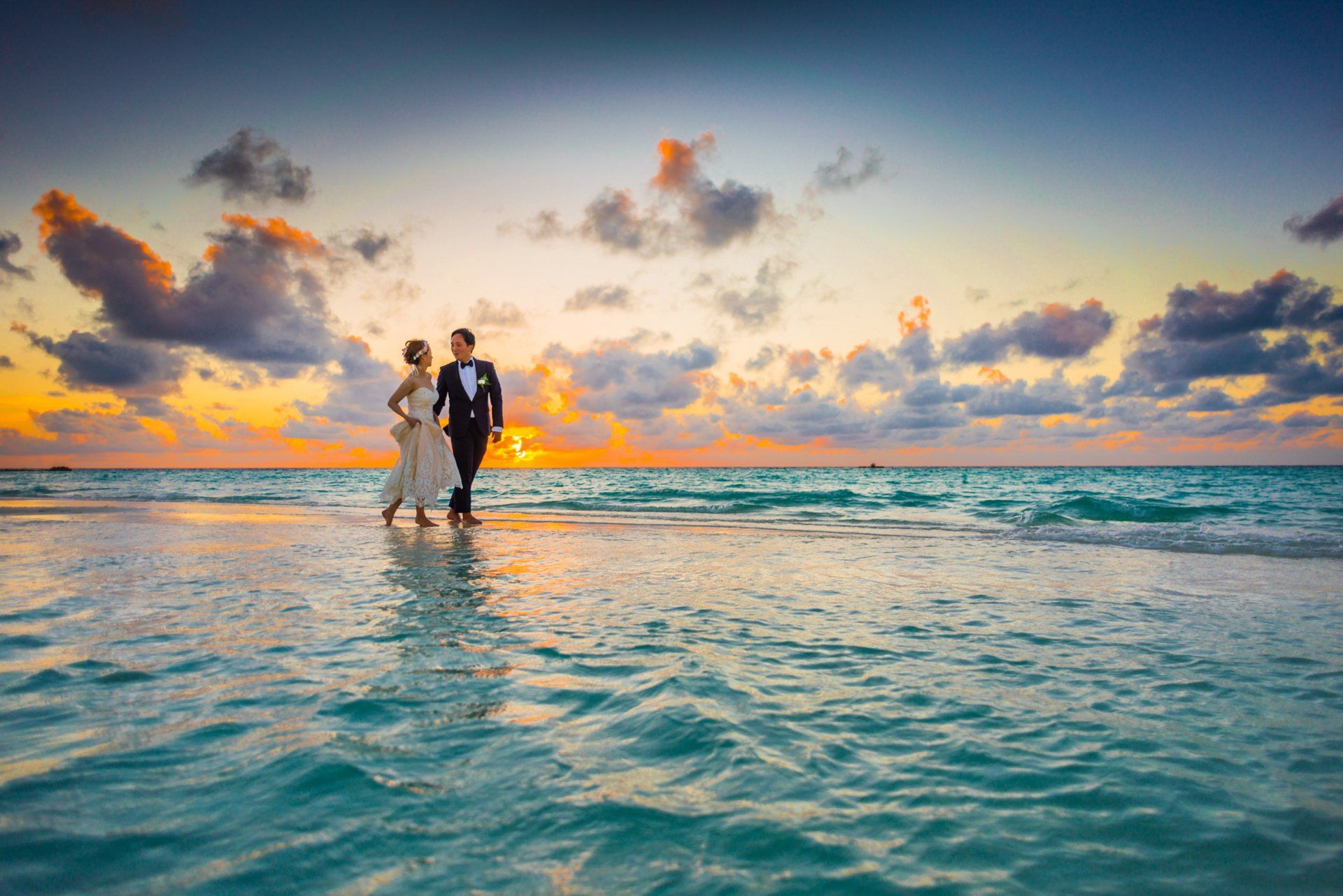 A bride and groom are walking on the beach at sunset.