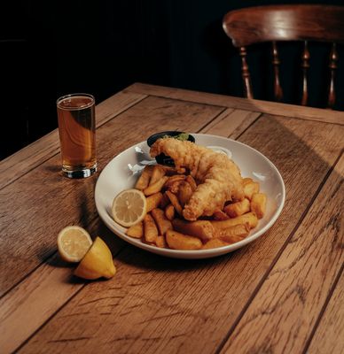 A plate of fish and chips sits on a wooden table next to a glass of beer