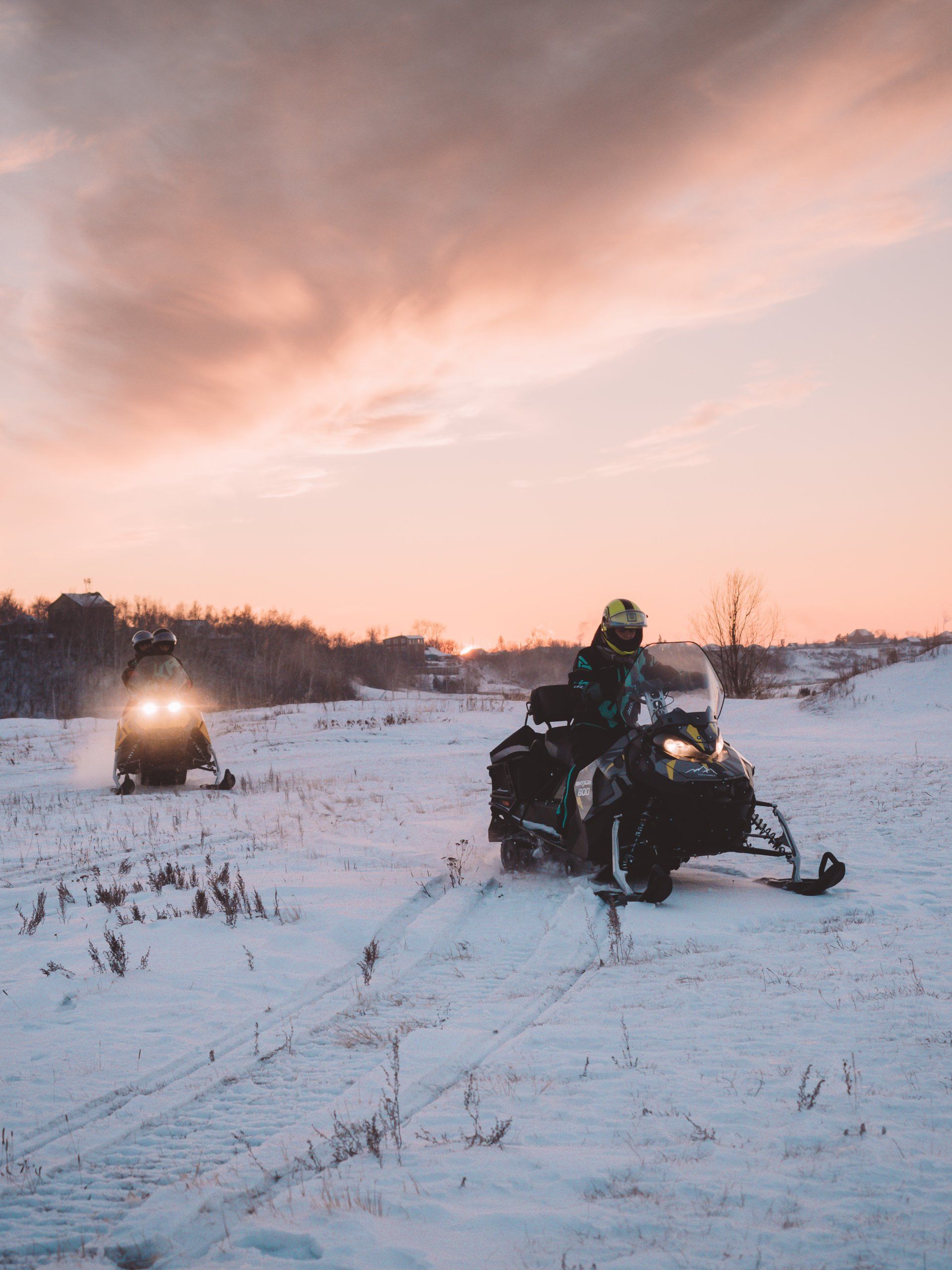 Three people snowmobiling through a field at sunset
