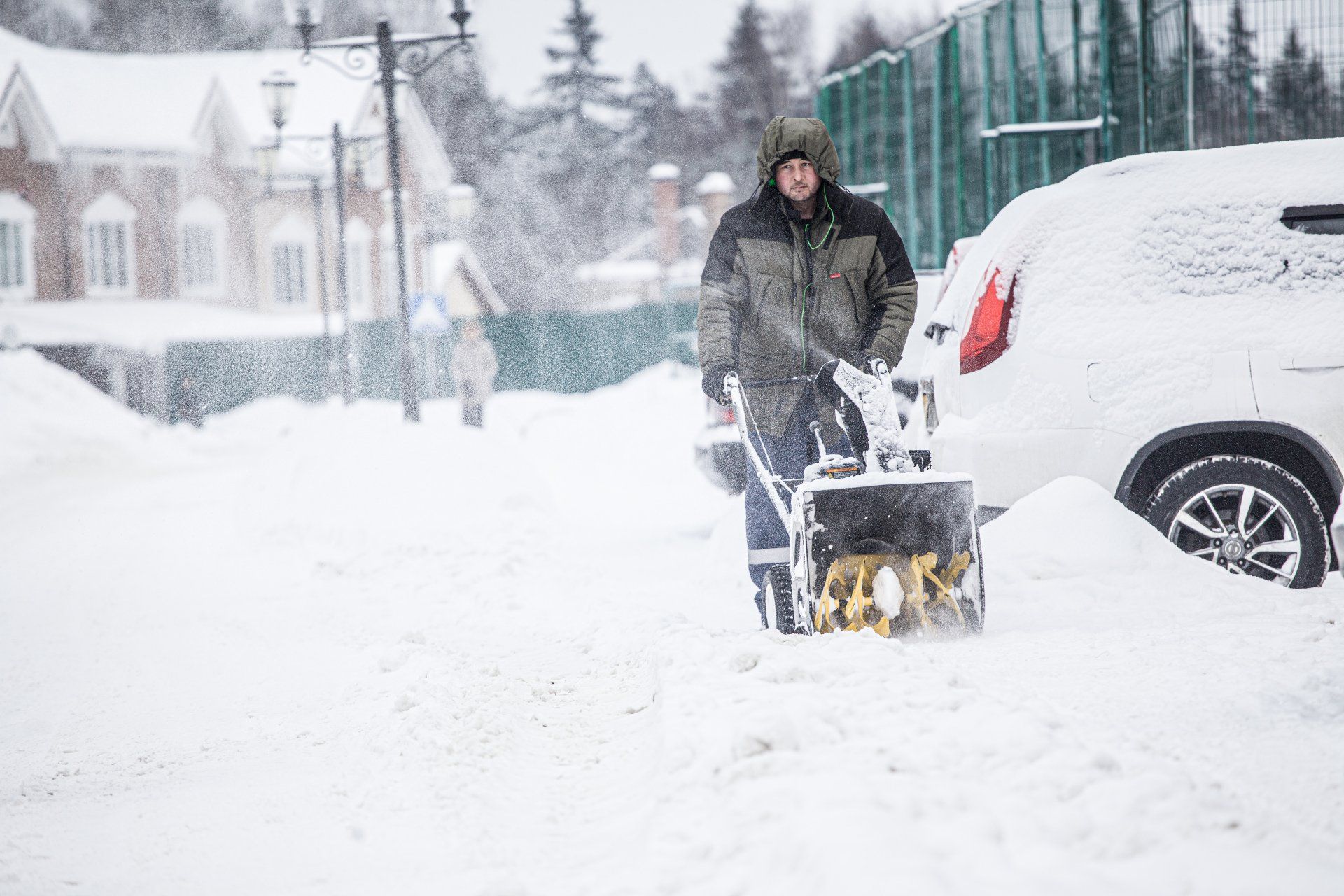 Man operating a snowblower on a snowy street, clearing snow near a parked white car and houses.