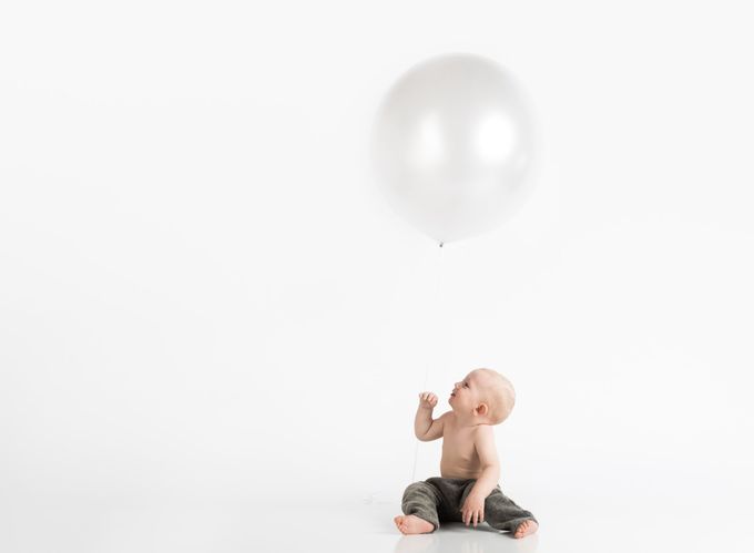 A baby is sitting on the floor looking up at a white balloon.