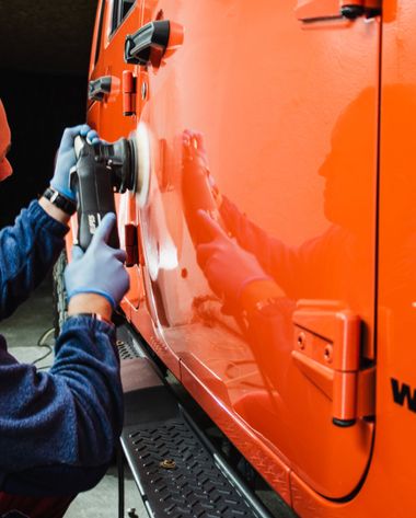 Person polishing an orange Jeep door with a power buffer, wearing gloves, in a garage.