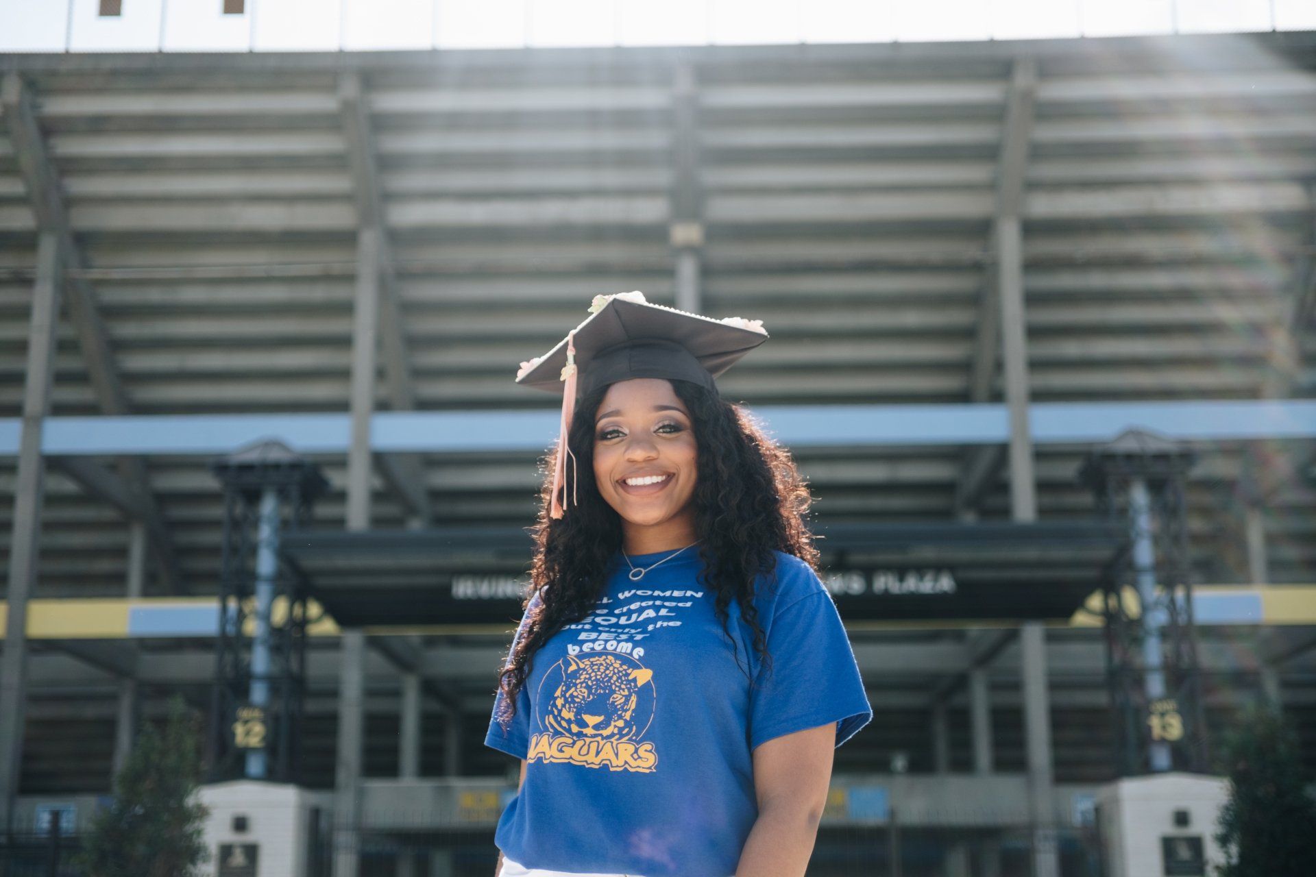 A woman wearing a graduation cap and gown is standing in front of a stadium.