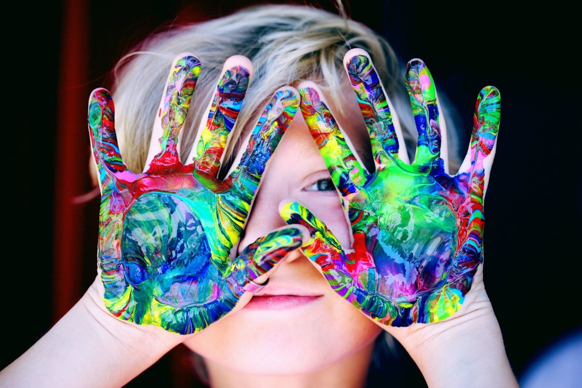 A young boy is covering his eyes with his hands painted in different colors.