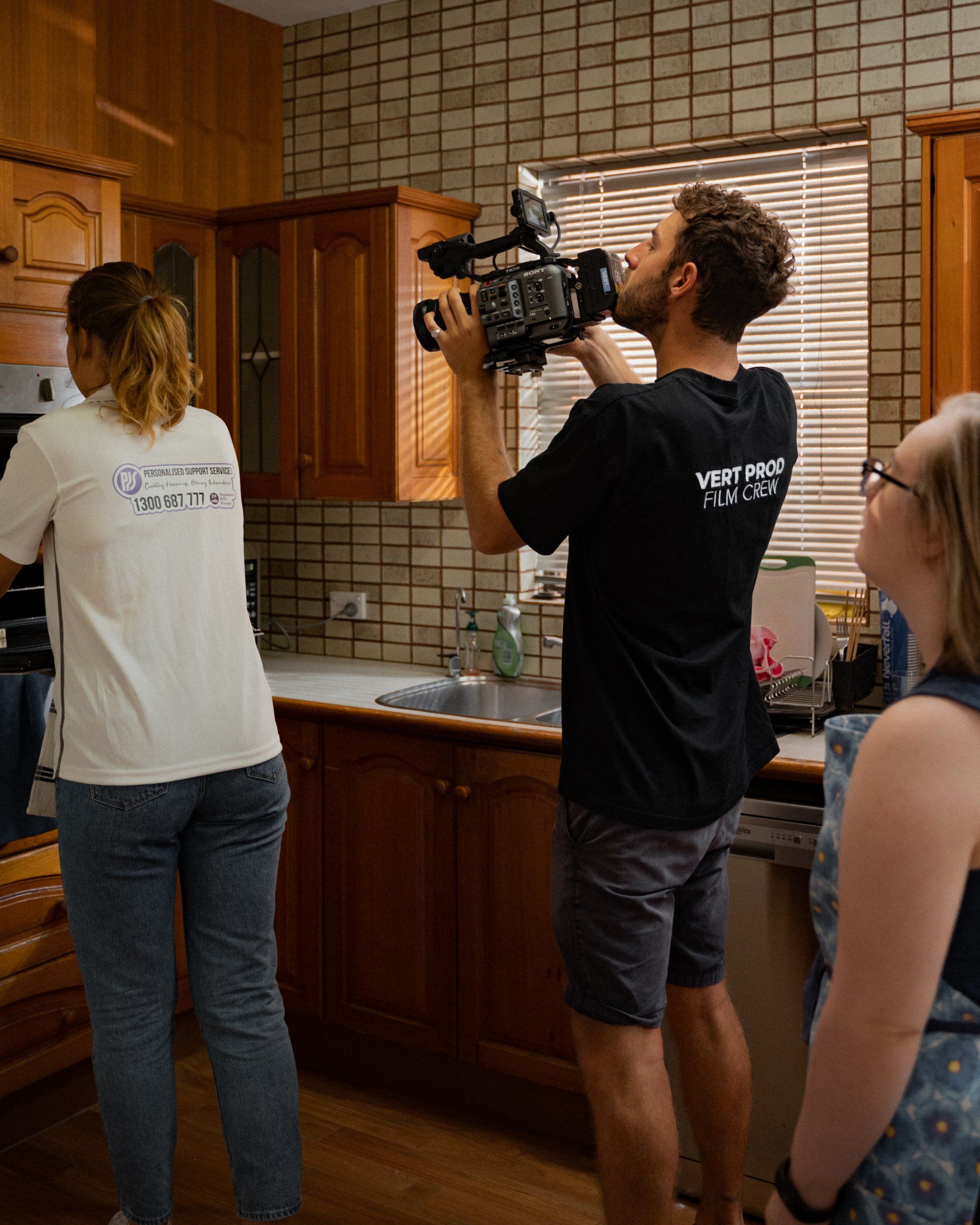 A man is holding a camera in a kitchen while a woman looks on